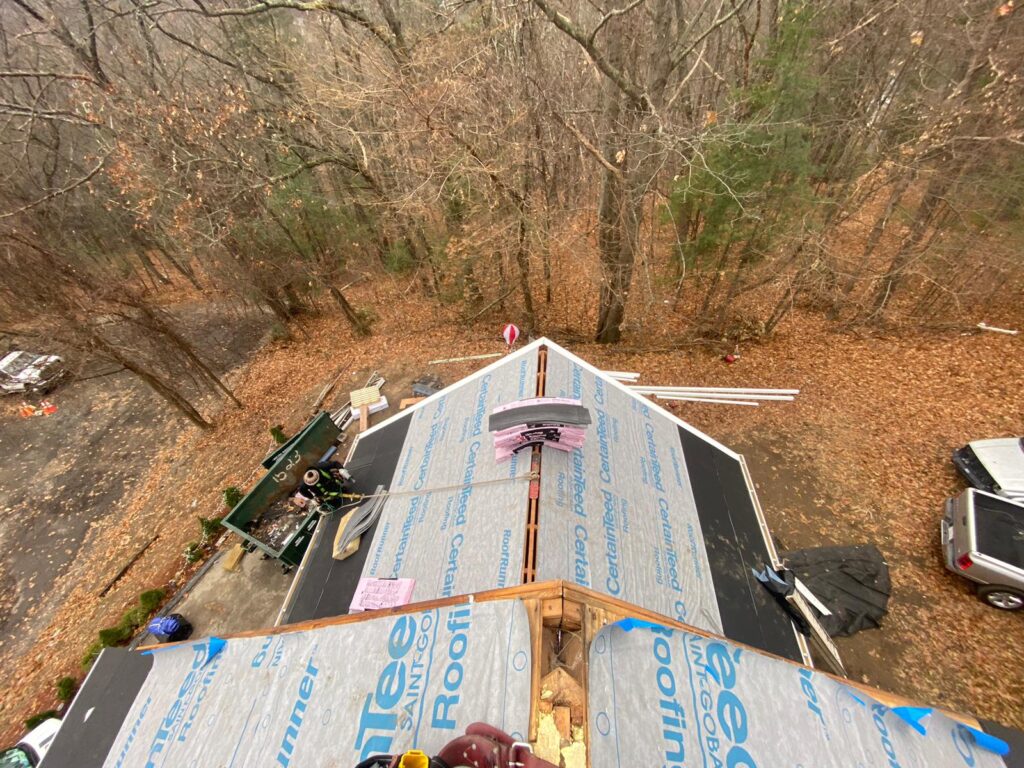 Aerial view of a roof replacement in Methuen, MA, showing workers on the roof with protective gear. The roof is covered with underlayment material, and the surrounding area has a dumpster for debris disposal.