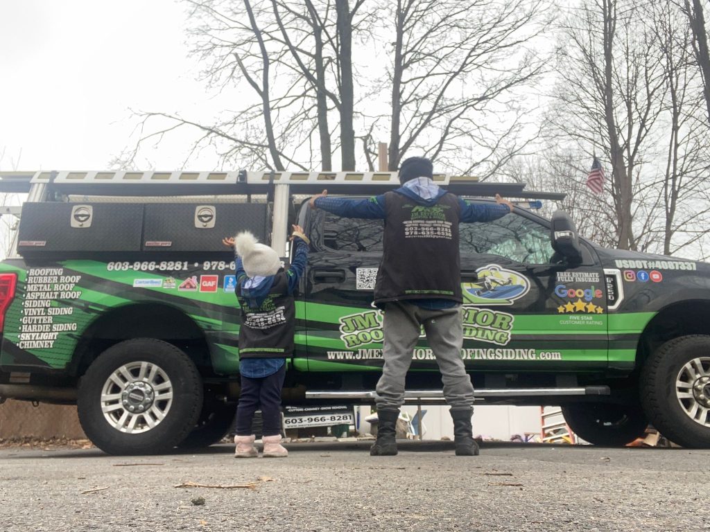 JM Exteriors Roofing Siding professional work truck in Hudson, NH, featuring a team member and child in company vests. The vehicle displays a 5-star Google rating and lists services including roof replacement, vinyl siding, and gutter installation.