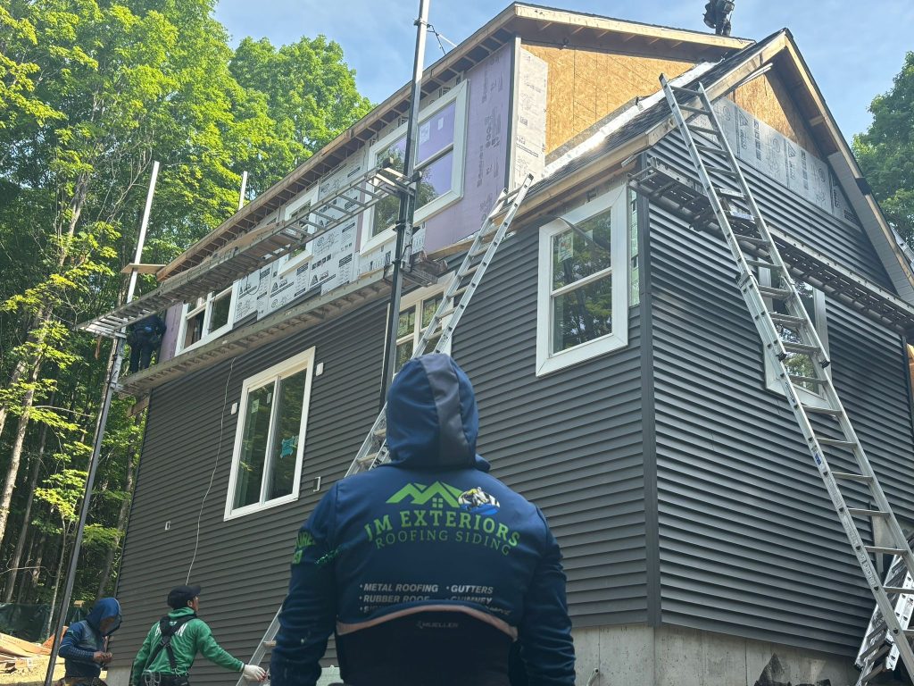 A JM Exteriors Roofing Siding team member in uniform supervises a siding installation at a two-story residential home in Londonderry, NH, with scaffolding and ladders in place.
